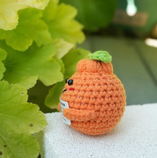 Crocheted orange pear with a green leaf on top, placed on a white surface with green leaves in the background.