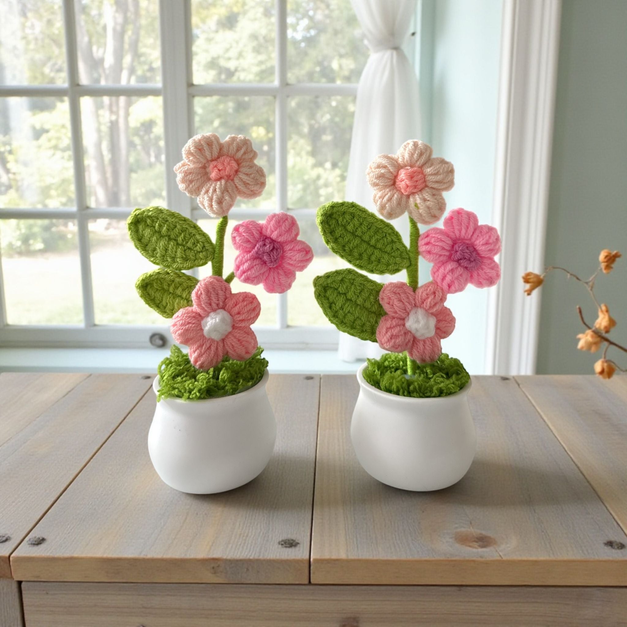 Two crocheted flower arrangements in white pots on a wooden table with a window in the background.