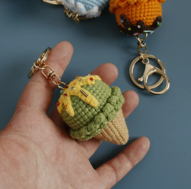 Hand holding a green and yellow crocheted keychain with other keychains in the background.