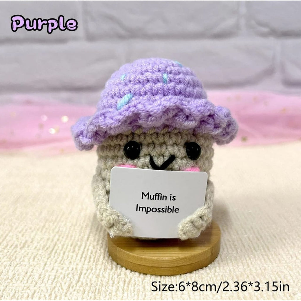 Crocheted character with a purple hat holding a sign that says 'Muffin is Impossible' on a soft background.