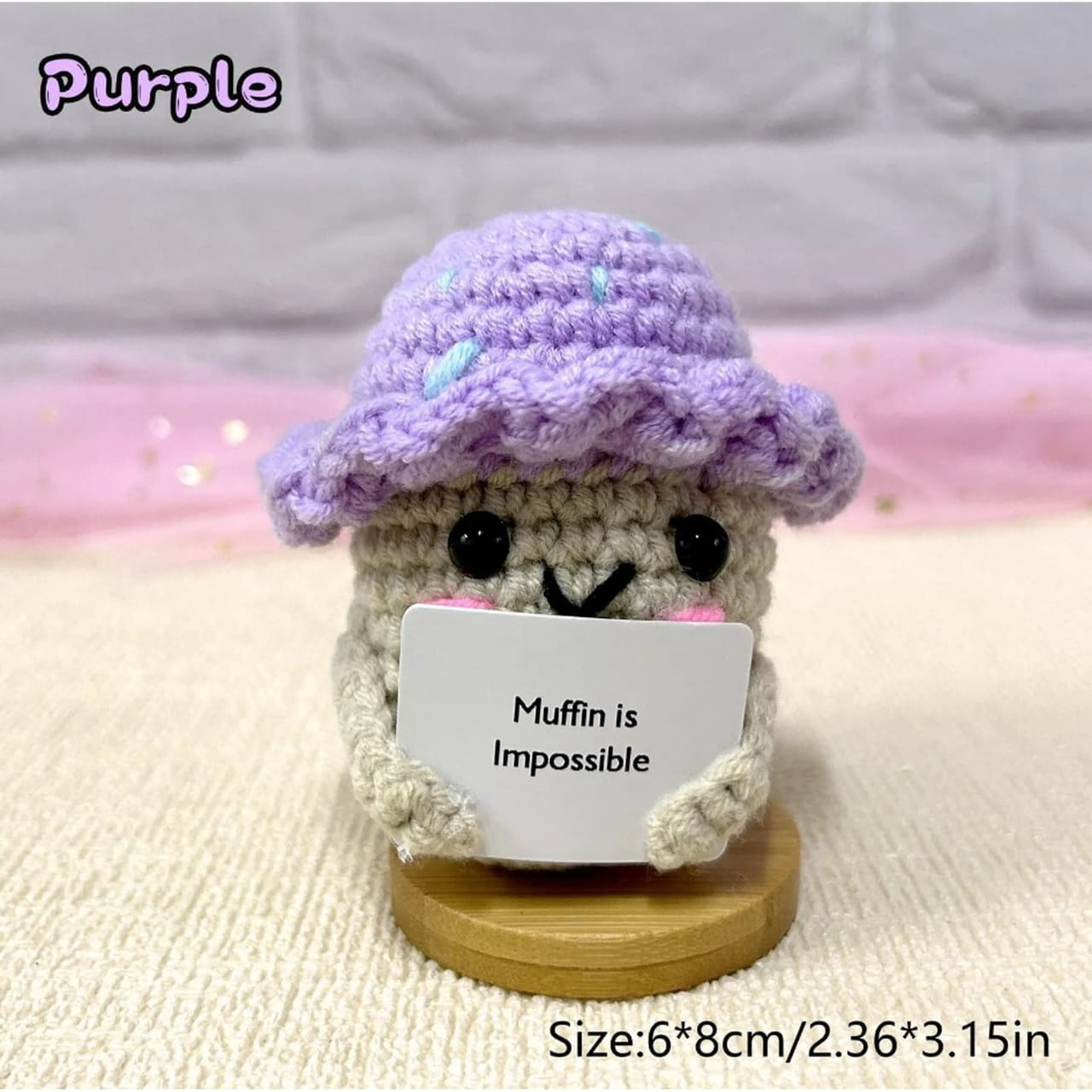 Crocheted character with a purple hat holding a sign that says 'Muffin is Impossible' on a soft background.