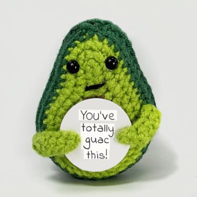 Crocheted avocado plushie holding a sign with text on a white background