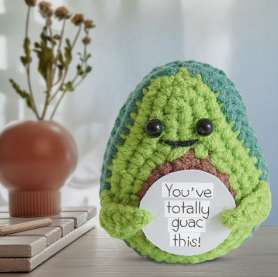 Crocheted avocado plush toy with a humorous message on a blurred background