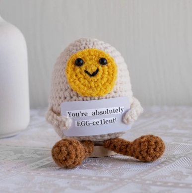 Crocheted egg-shaped character with a smiling face and a tag saying 'You're absolutely EGG-cellent!' on a neutral background.