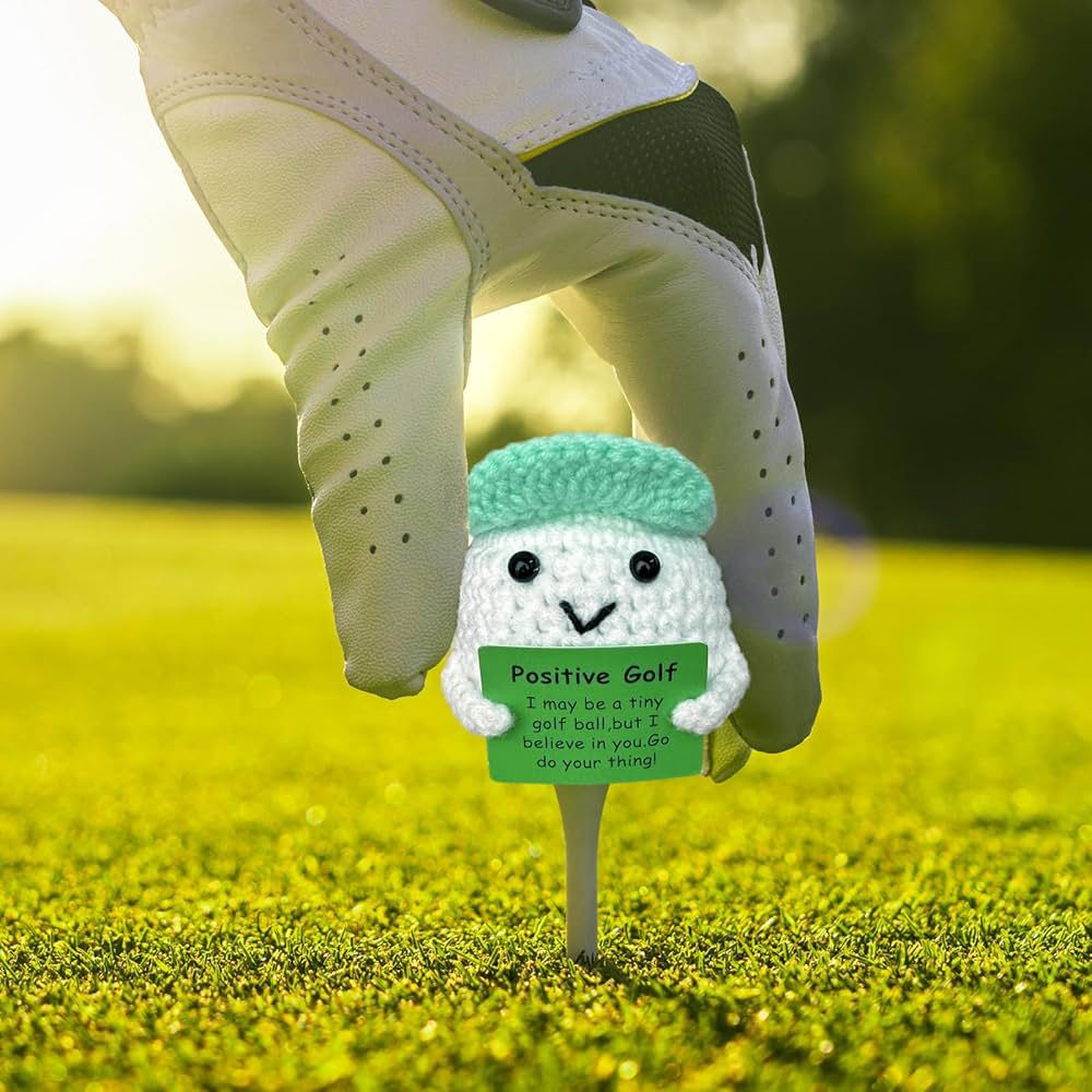 Golf tee with a small plush toy character on a golf course, wearing a green hat and holding a sign.