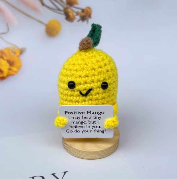 Crocheted yellow mango character holding a card with text on a white background