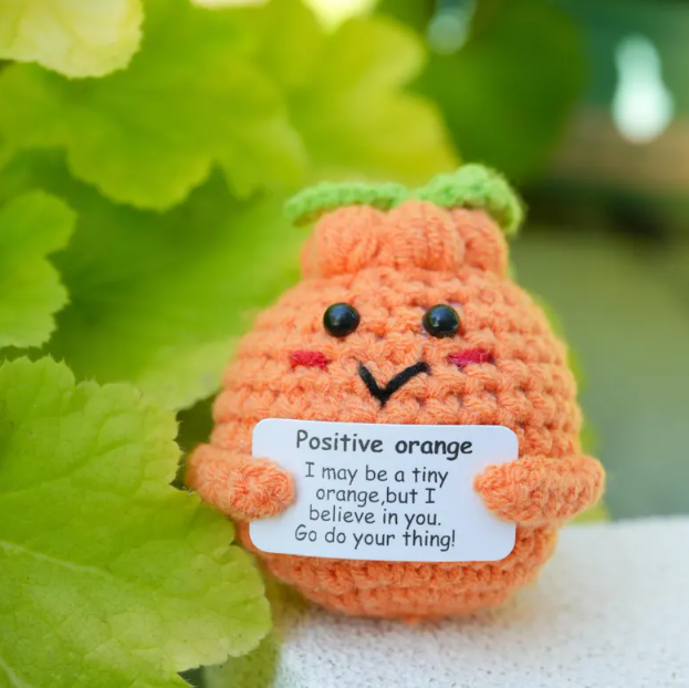 Crocheted orange character with a message tag on green leaves