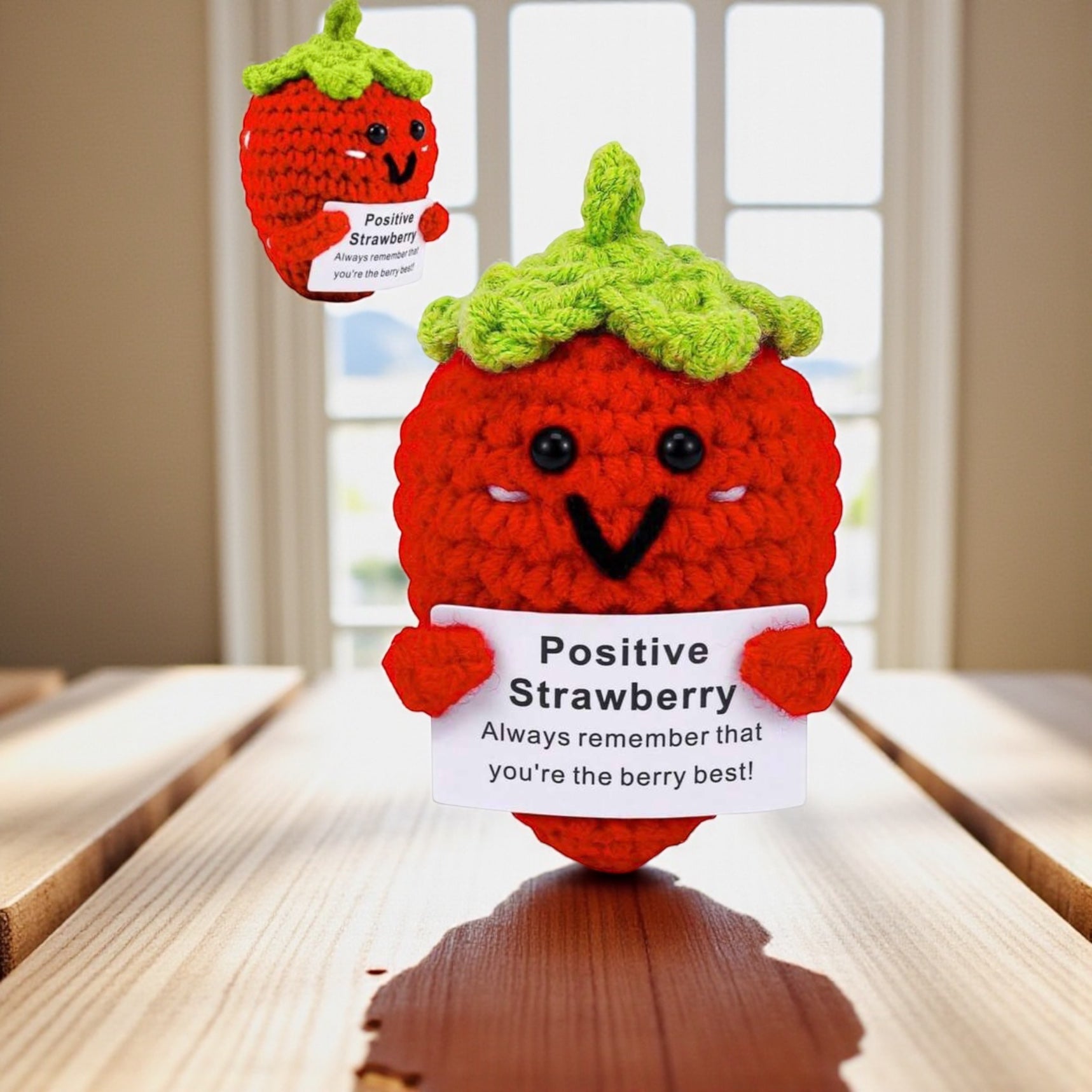 Crocheted strawberry plush toy holding a sign with motivational message on a wooden surface.
