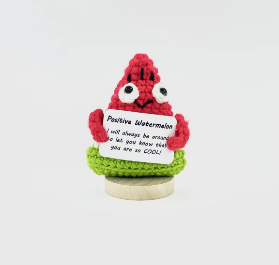 Crocheted watermelon character holding a sign with text on a white background