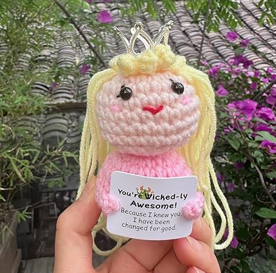 Crocheted doll with blonde hair and a pink outfit, held in front of a garden background.