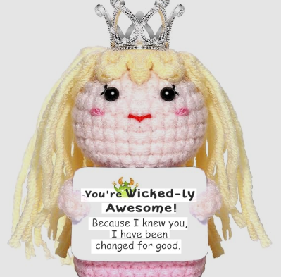 Crocheted doll with blonde hair, pink dress, and tiara holding a sign saying 'You're Wicked-ly Awesome!'.