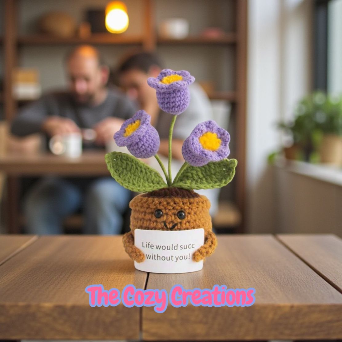 Crocheted flower pot with flowers and a small bear holding a sign, set on a wooden table with a blurred background of people and shelves.