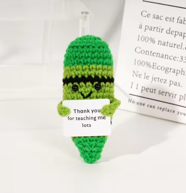Green crocheted bottle cover with a smiley face holding a card, on a white background.
